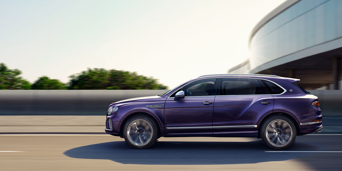 Bentley Jeddah Bentley Bentayga Extended Wheelbase Mulliner luxury SUV profile in Tanzanite Purple paint, driving on an country road past trees.