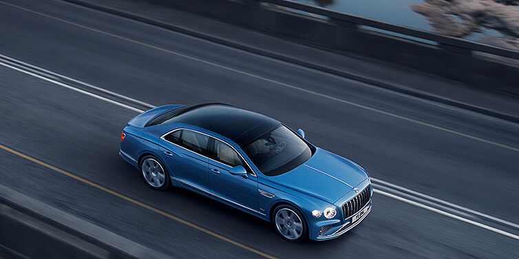 Bentley Jeddah New Bentley Flying Spur Azure sedan in Light windsor blue paint by Mulliner, with 22 inch Azure wheels seen from overhead driving dynamically on a country road