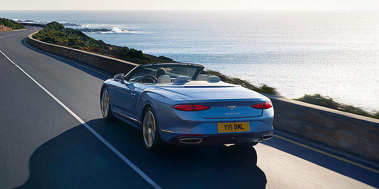 Bentley Jeddah New Bentley Continental GTC Azure convertible rear view in Light Windsor blue paint with 22 inch ten spoke wheels driving dynamically on an ocean road in the sunshine